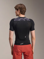 Alpinestars A-Motion Plasma Protection Jacket - Short Sleeve, kurtka ochronna z krótkim rękawem, czarna z szarymi akcentami kamuflażu na górnej części pleców, widok z tyłu prezentujący zintegrowany ochraniacz pleców i białe logo Astars na dole, zestawiona z czerwonymi szortami, techniczna odzież ochronna MTB