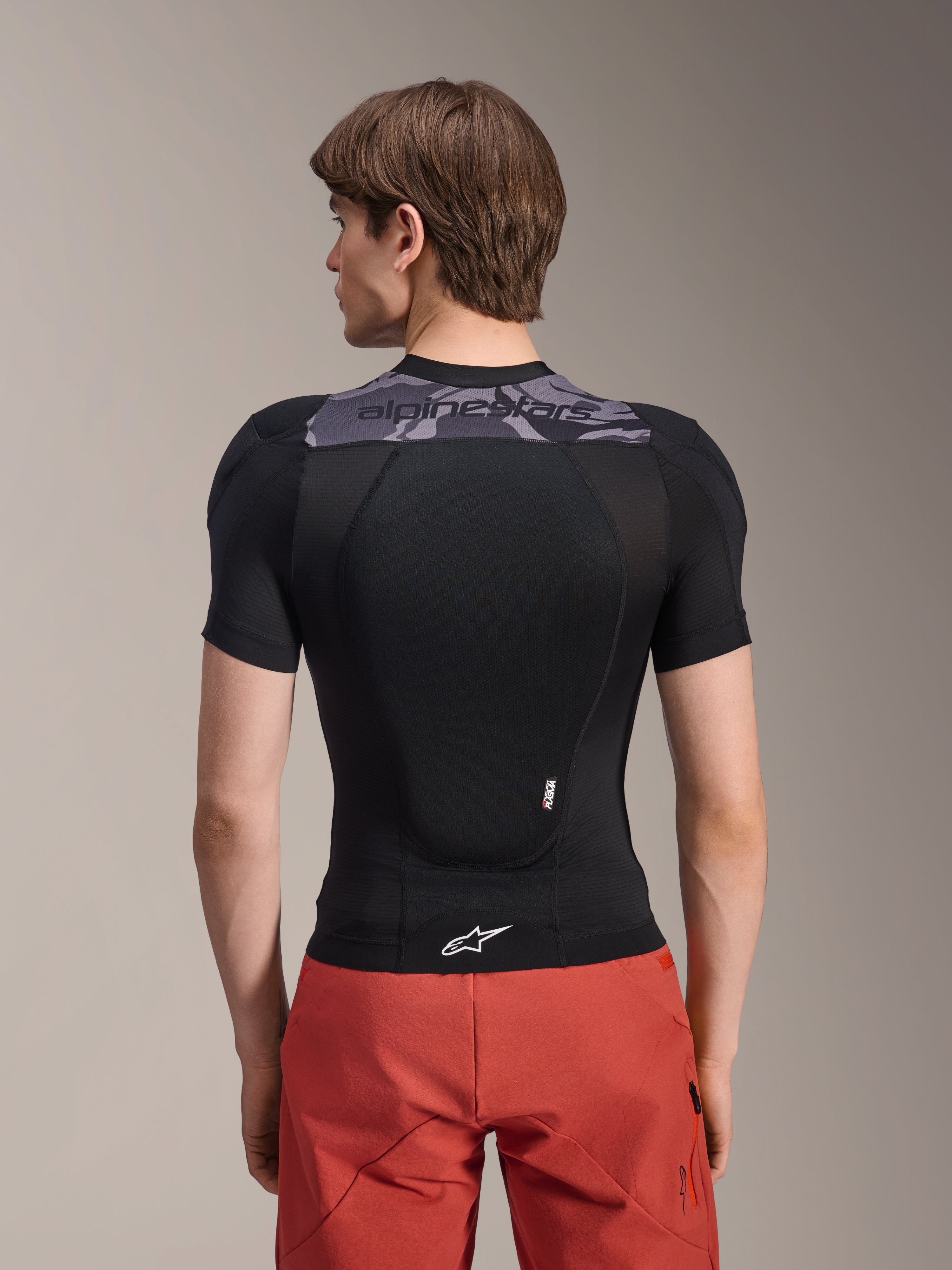 Alpinestars A-Motion Plasma Protection Jacket - Short Sleeve, kurtka ochronna z krótkim rękawem, czarna z szarymi akcentami kamuflażu na górnej części pleców, widok z tyłu prezentujący zintegrowany ochraniacz pleców i białe logo Astars na dole, zestawiona z czerwonymi szortami, techniczna odzież ochronna MTB