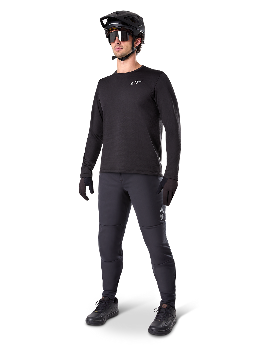 Rowerzysta górski w spodniach Alpinestars A-Dura Thermal, czarne, zestawione z czarną koszulką z długim rękawem, czarnym kaskiem i okularami przeciwsłonecznymi, srebrne logo Alpinestars na lewym udzie, zaprojektowane do jazdy na szlaku w zimne dni