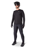 Rowerzysta górski w spodniach Alpinestars A-Dura Thermal, czarne, zestawione z czarną koszulką z długim rękawem, czarnym kaskiem i okularami przeciwsłonecznymi, srebrne logo Alpinestars na lewym udzie, zaprojektowane do jazdy na szlaku w zimne dni