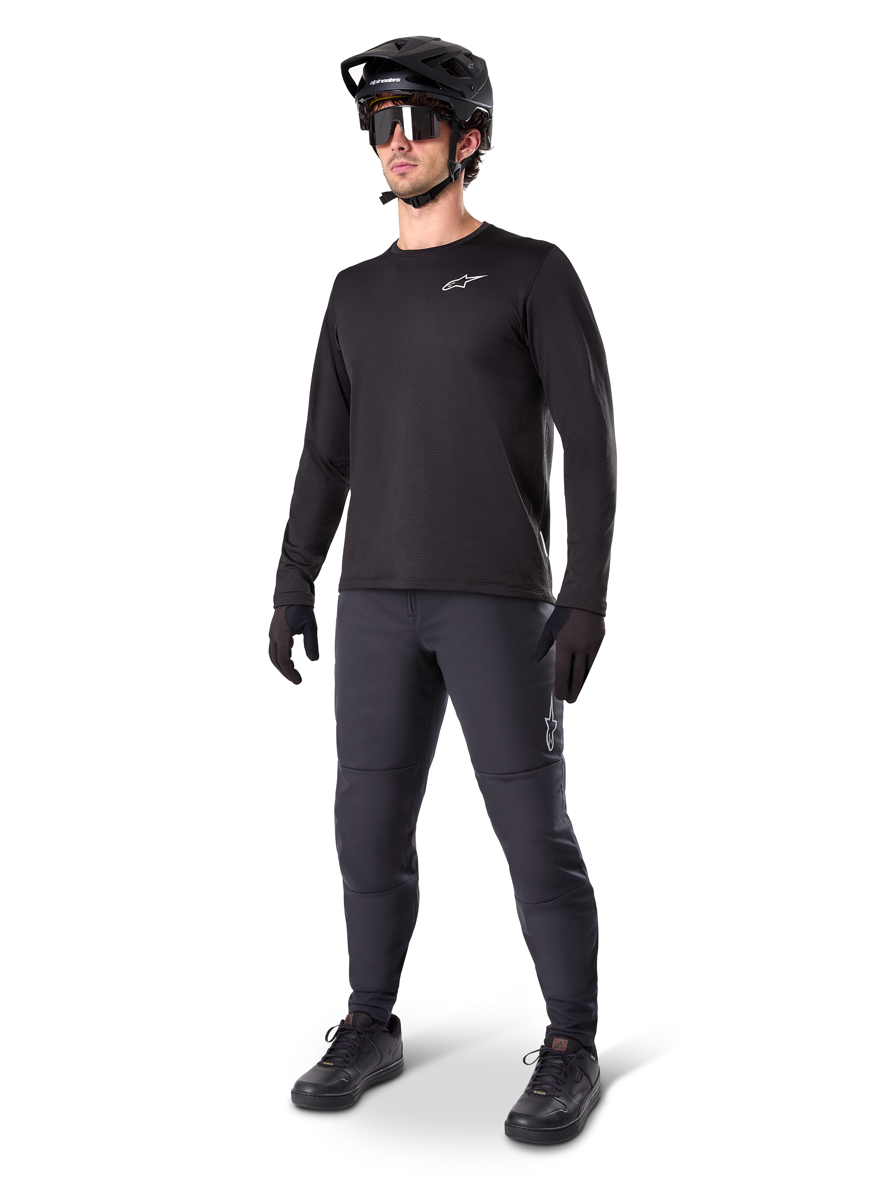 Rowerzysta górski w spodniach Alpinestars A-Dura Thermal, czarne, zestawione z czarną koszulką z długim rękawem, czarnym kaskiem i okularami przeciwsłonecznymi, srebrne logo Alpinestars na lewym udzie, zaprojektowane do jazdy na szlaku w zimne dni