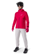 Model ubrany w spodnie Alpinestars A-Dura Waterproof w kolorze Titanium gray, zestawione z jaskrawoczerwoną wodoodporną kurtką i pasującymi rękawicami, czarny kask MTB z goglami oraz czarne buty, kompletny strój na rower górski na mokre warunki na szlaku