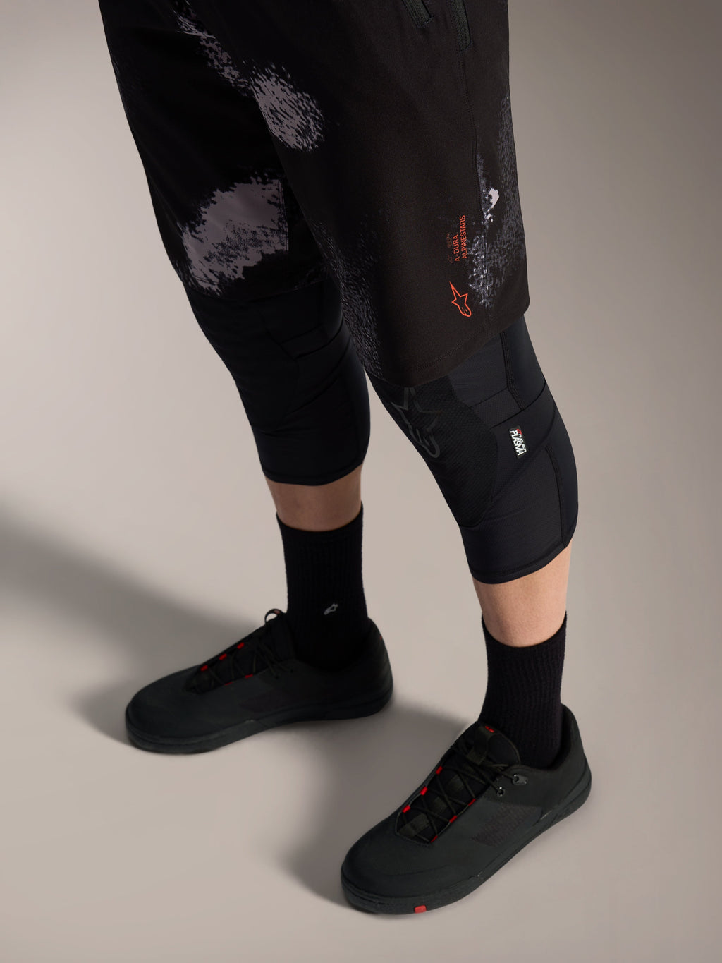 Szorty Alpinestars A-Dura Ride Black Camo z pomarańczowym logo, noszone z ochraniaczami kolan i butami MTB.