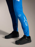 Spodnie Alpinestars A-Supra Elite Faded Bright Blue, białe logo Astars na dolnej części nogawki i czarne buty MTB.