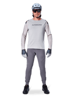 Alpinestars A-Aria Elite Pants, ciemnoszare spodnie rowerowe MTB, noszone przez rowerzystę w jasnoszarej koszulce z długim rękawem, jasnoniebieskim kasku i czarnych butach, z widocznymi laserowymi perforacjami na wewnętrznej stronie nogawek i dopasowanym sportowym krojem.