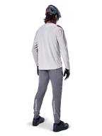 Alpinestars A-Aria Elite Pants, spodnie rowerowe MTB, ciemnoszare, widok z tyłu na rowerzystę w pełnym stroju, z białym logo Alpinestars na dolnej części nogawek, sportowy krój z laserowymi perforacjami, zestawione z jasnoszarą koszulką z długim rękawem i szarym kaskiem MTB