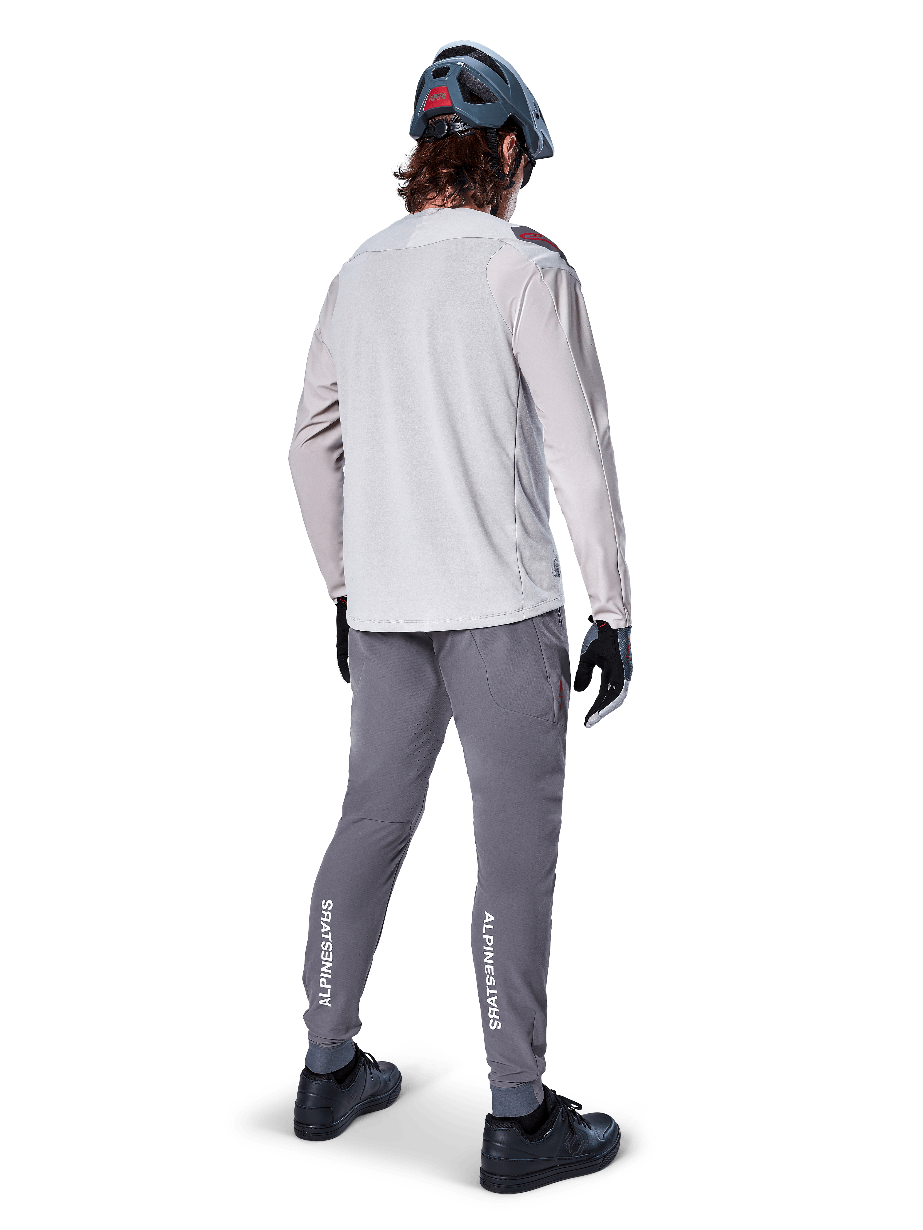 Alpinestars A-Aria Elite Pants, spodnie rowerowe MTB, ciemnoszare, widok z tyłu na rowerzystę w pełnym stroju, z białym logo Alpinestars na dolnej części nogawek, sportowy krój z laserowymi perforacjami, zestawione z jasnoszarą koszulką z długim rękawem i szarym kaskiem MTB