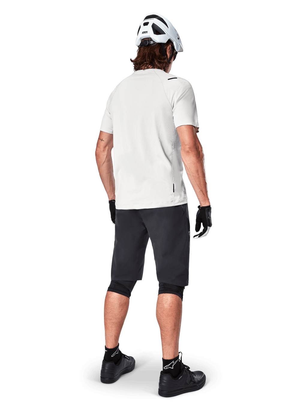Rowerzysta górski widoczny od tyłu, ubrany w szorty Alpinestars A-Aria w kolorze czarnym, białą techniczną koszulkę z krótkim rękawem, biały kask, czarne rękawiczki i czarne buty MTB, odzież rowerowa do jazdy po szlakach