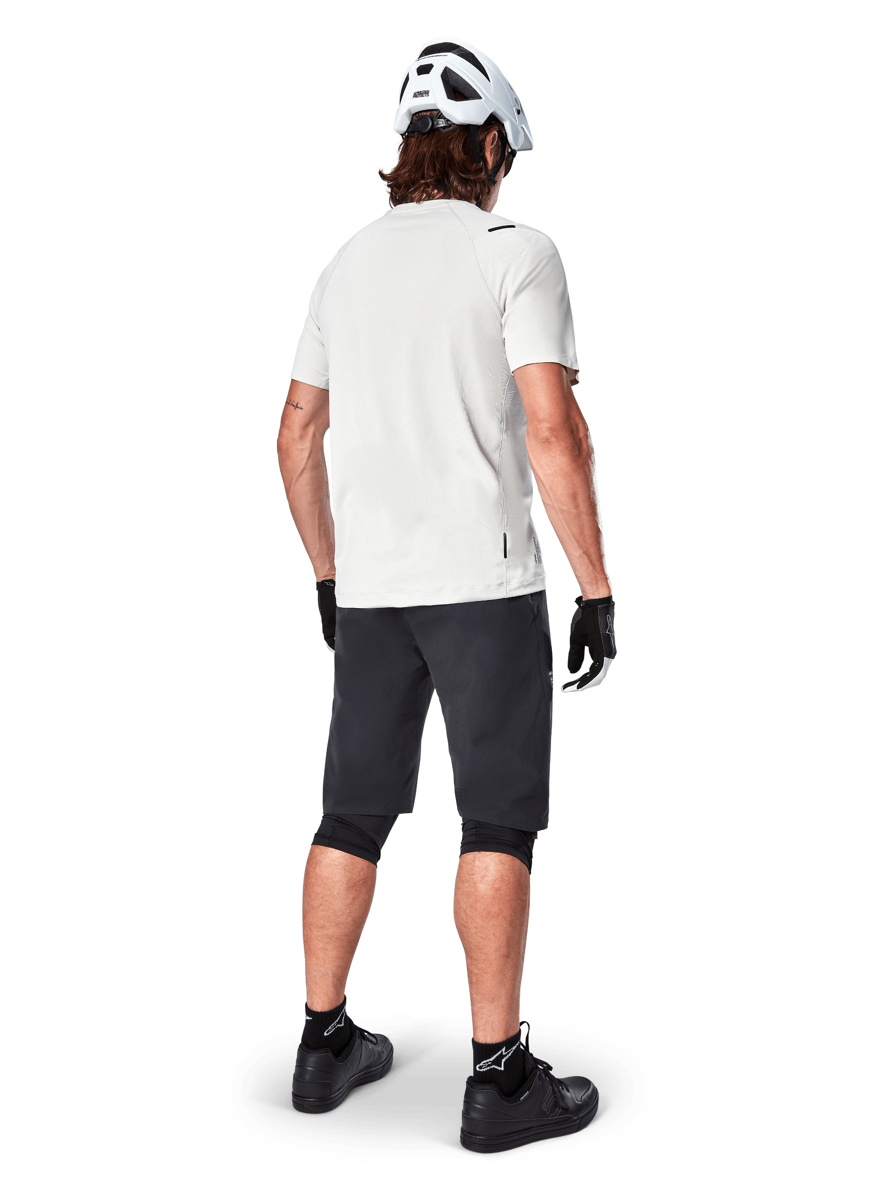 Rowerzysta górski widoczny od tyłu, ubrany w szorty Alpinestars A-Aria w kolorze czarnym, białą techniczną koszulkę z krótkim rękawem, biały kask, czarne rękawiczki i czarne buty MTB, odzież rowerowa do jazdy po szlakach