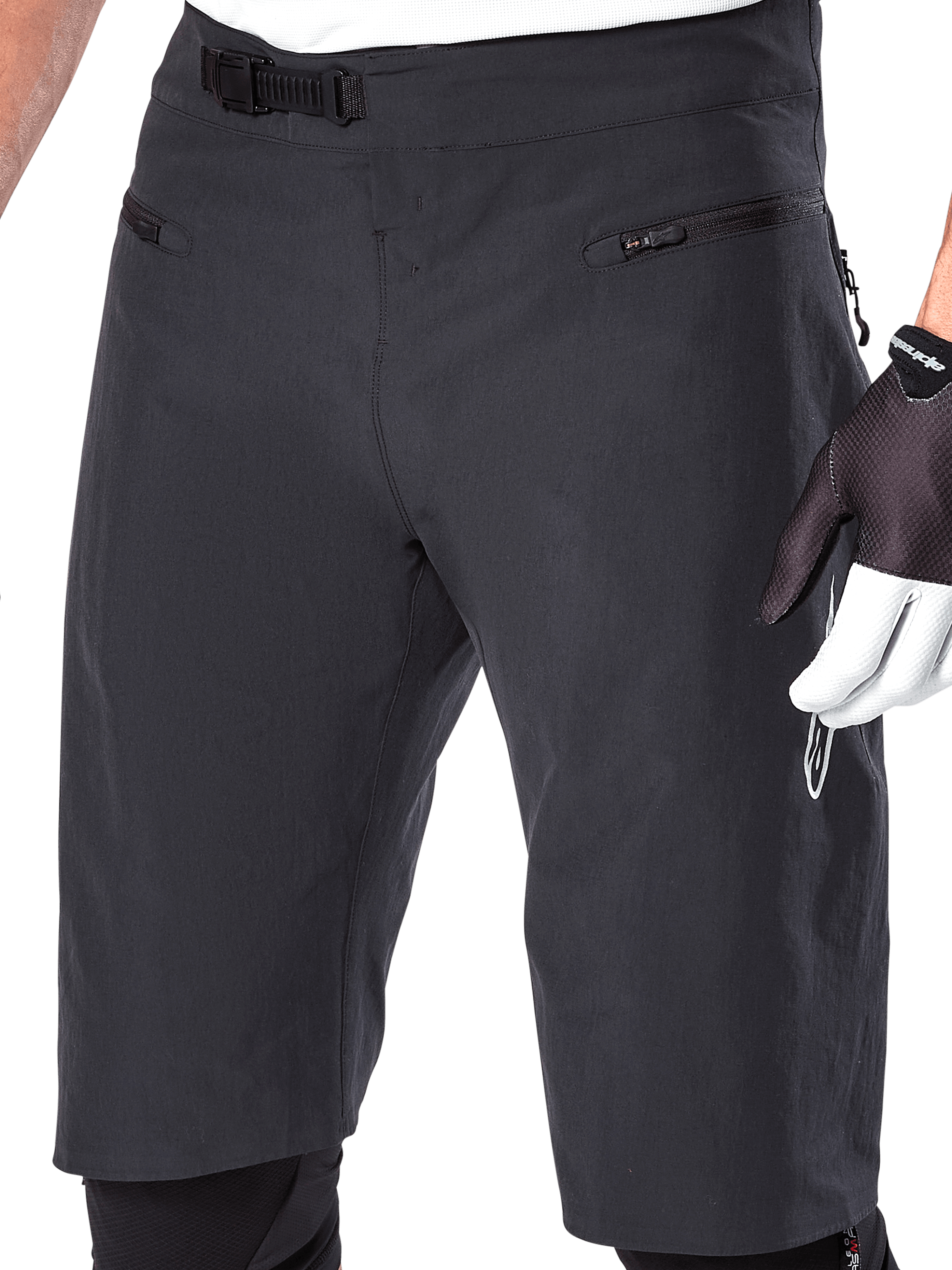 Alpinestars A-Aria Shorts, spodenki rowerowe MTB, czarne, zbliżenie na przód pasa i uda pokazujące system zapięcia zapadkowego oraz kieszenie na zamek, noszone z czarno-białymi rękawiczkami rowerowymi, profesjonalna odzież trailowa