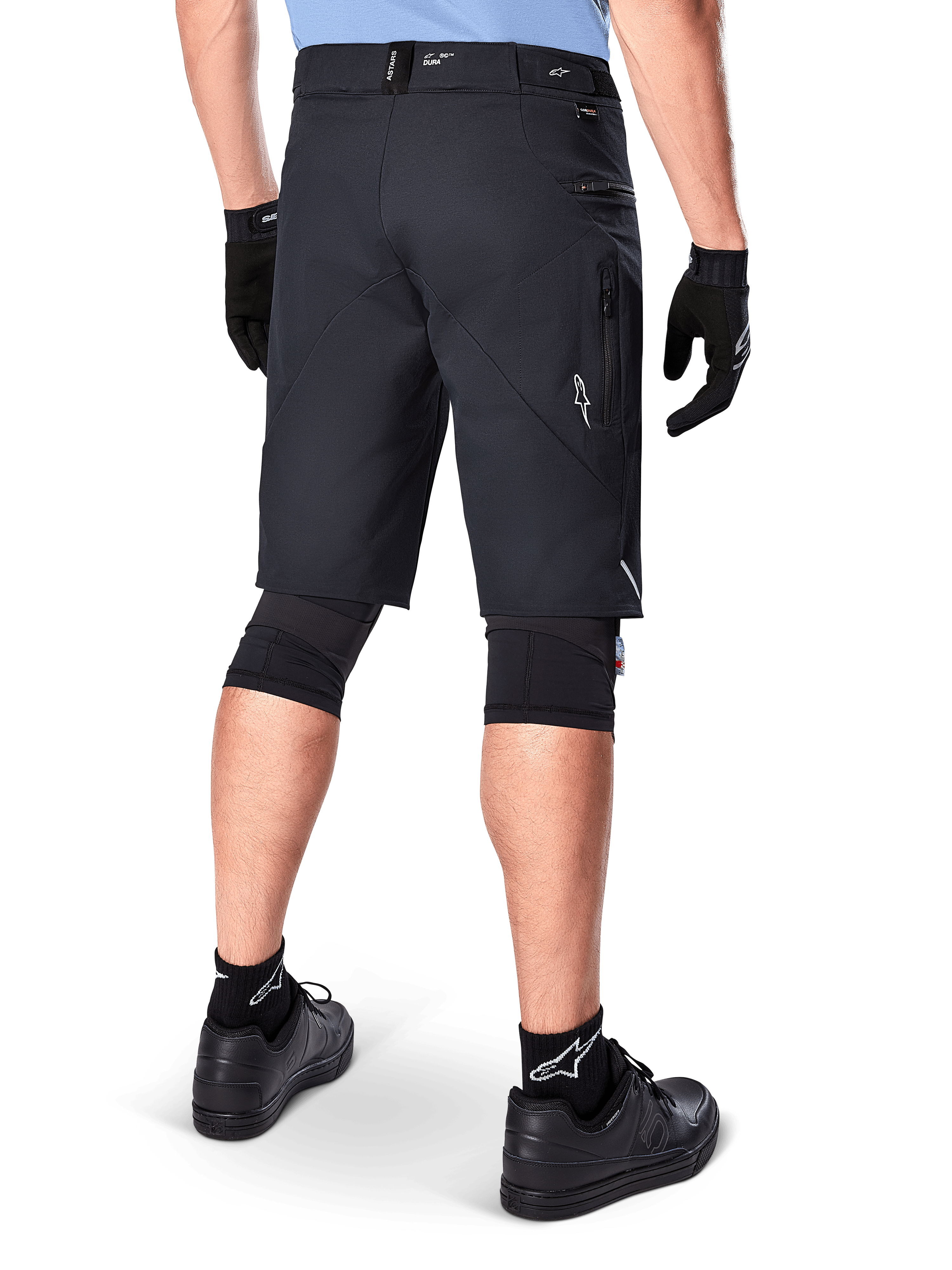 Alpinestars A-Dura Elite Shorts, czarne, widok od tyłu rowerzysty w stroju MTB, w tym czarne nakolanniki, rękawiczki i skarpetki z białym logo, boczne kieszenie na zamek i odblaskowe akcenty, wytrzymały materiał Cordura