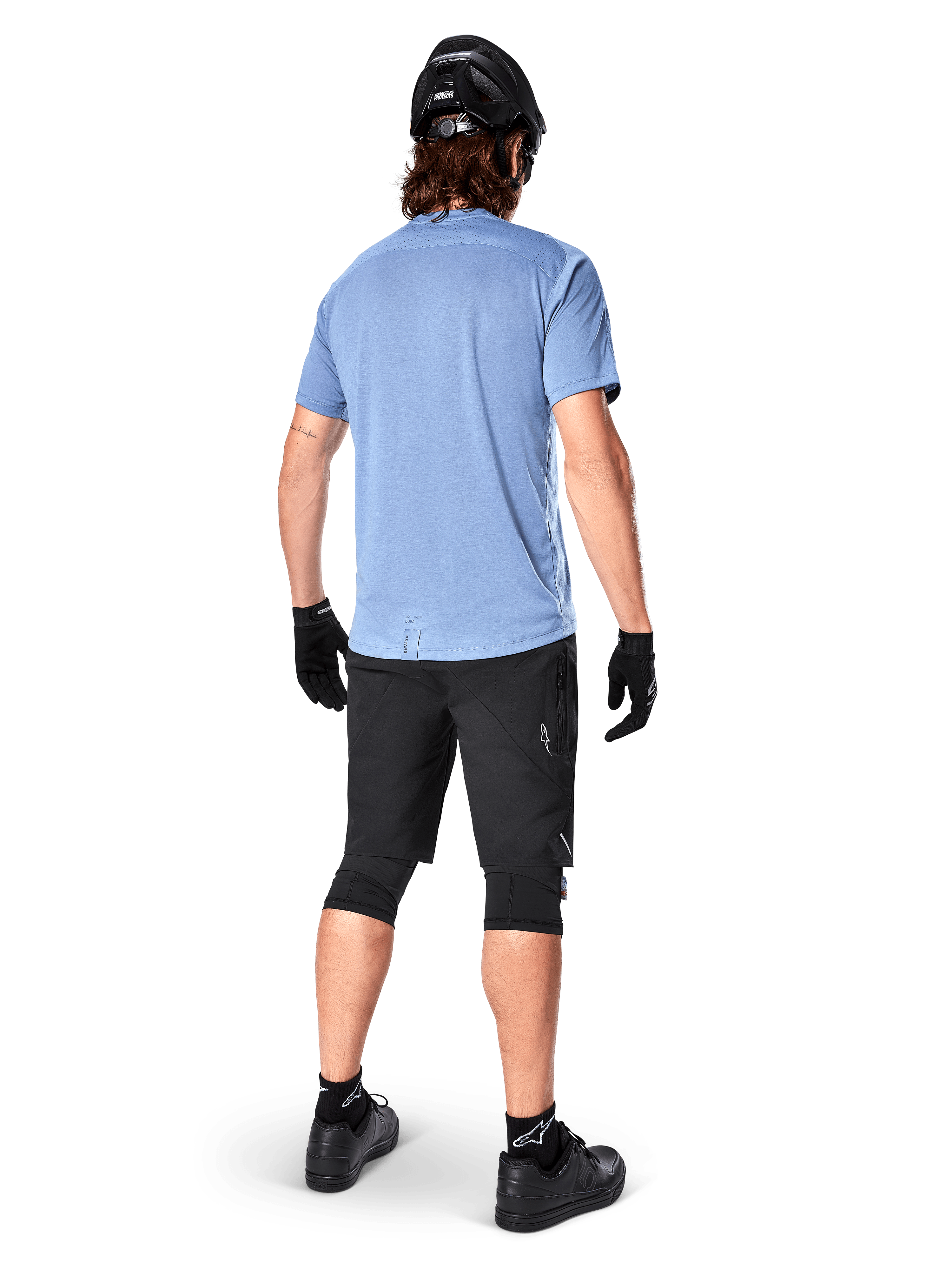 Alpinestars A-Dura Elite Shorts, spodenki rowerowe MTB, czarne, widok z tyłu na modelu, zestaw z jasnoniebieską koszulką z krótkim rękawem i czarnym sprzętem ochronnym, techniczna konstrukcja z kieszeniami na zamek i odblaskowymi detalami do jazdy w terenie