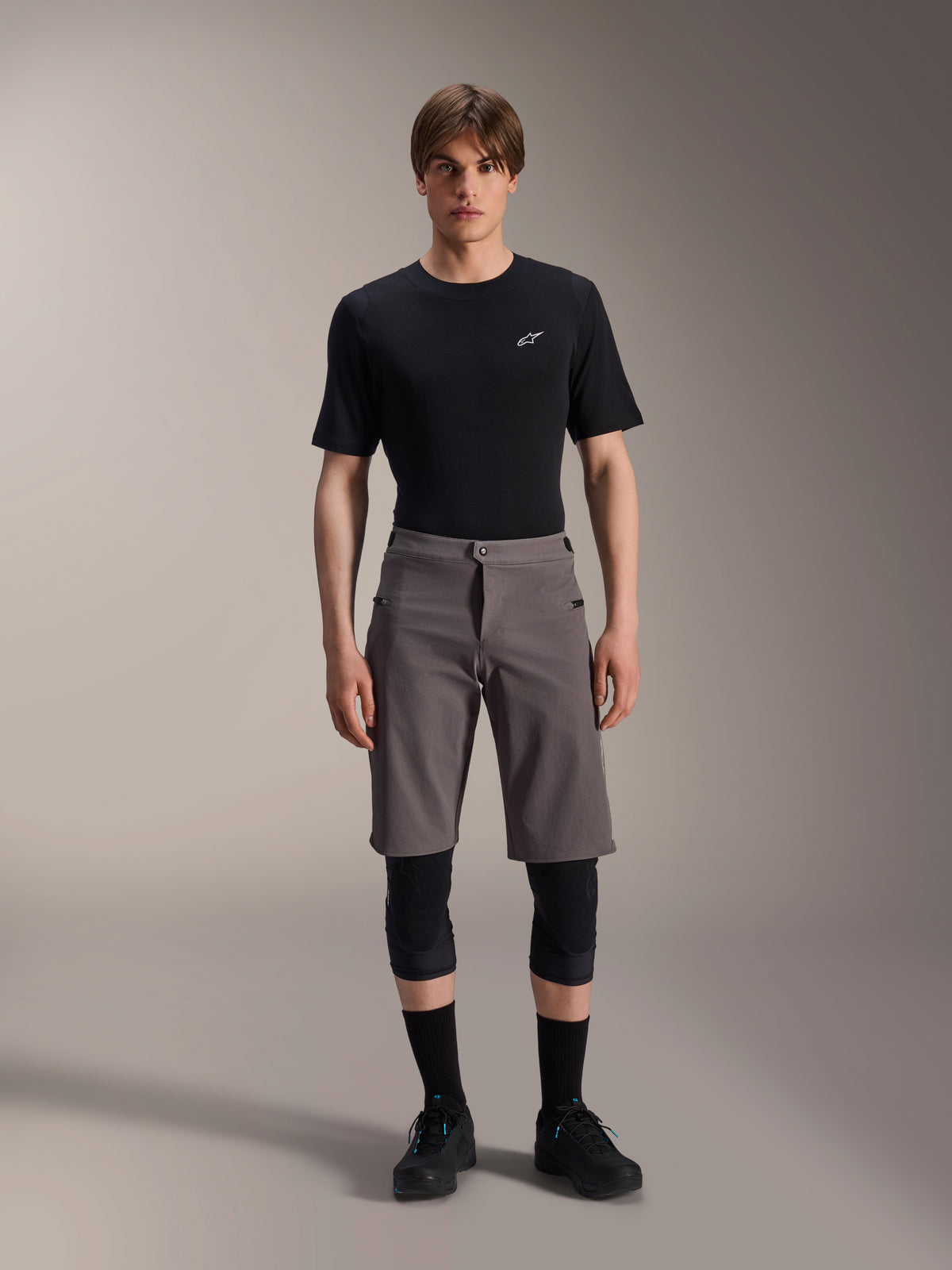 Alpinestars A-Dura Elite Shorts, ciemnoszare spodenki MTB, noszone z czarnym t-shirtem z okrągłym dekoltem i czarnymi ochraniaczami kolan, odzież rowerowa do kolan z zapięciem na zatrzask i wytrzymałym materiałem Cordura.