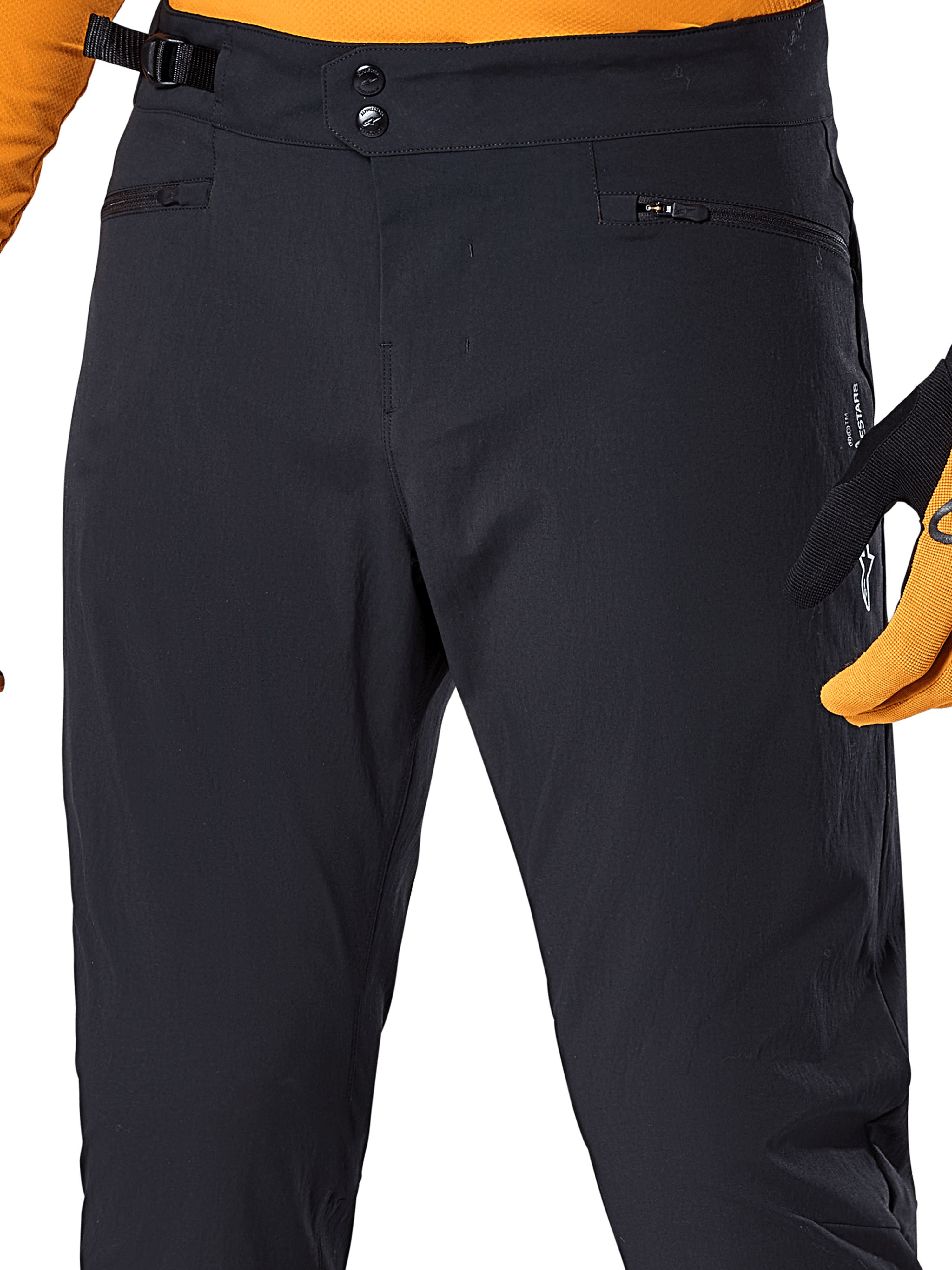 Alpinestars A-Dura Pants, czarne spodnie rowerowe MTB, zbliżenie na podwójne zapięcie na zatrzaski w pasie, boczną regulację dopasowania i kieszenie zapinane na zamek, zestawione z pomarańczową koszulką i rękawicami