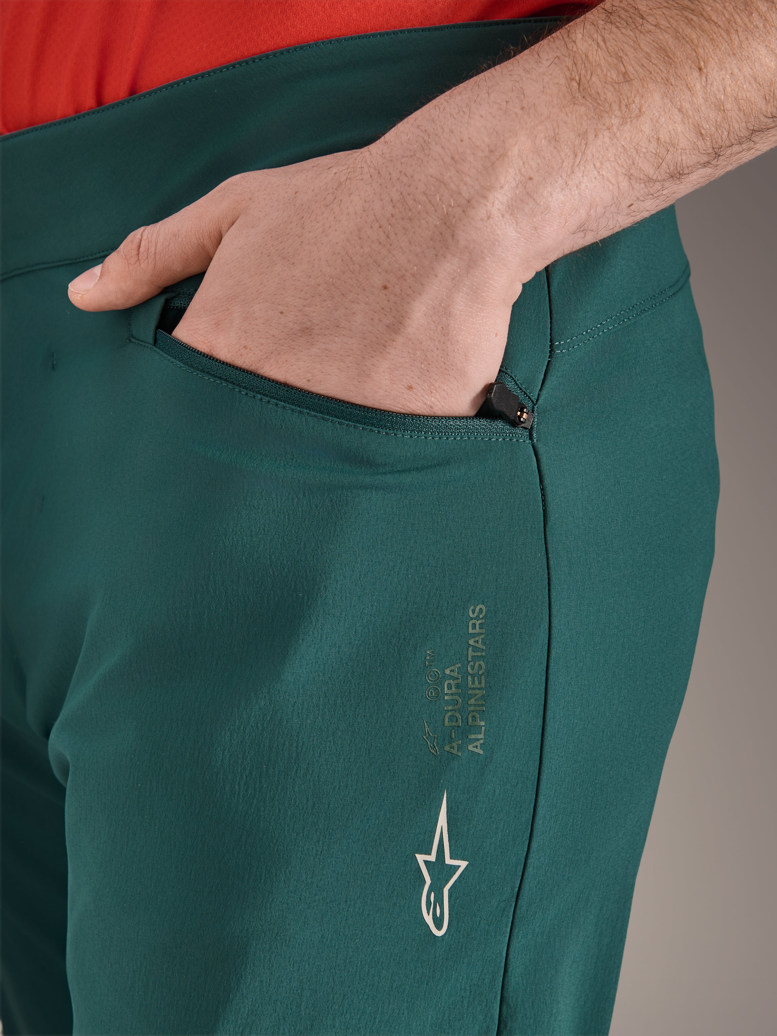 Spodnie Alpinestars A-Dura w kolorze Dark Pine, zbliżenie na kieszeń zapinaną na zamek z dłonią, białe logo i branding Alpinestars na udzie, wytrzymałe spodnie rowerowe MTB