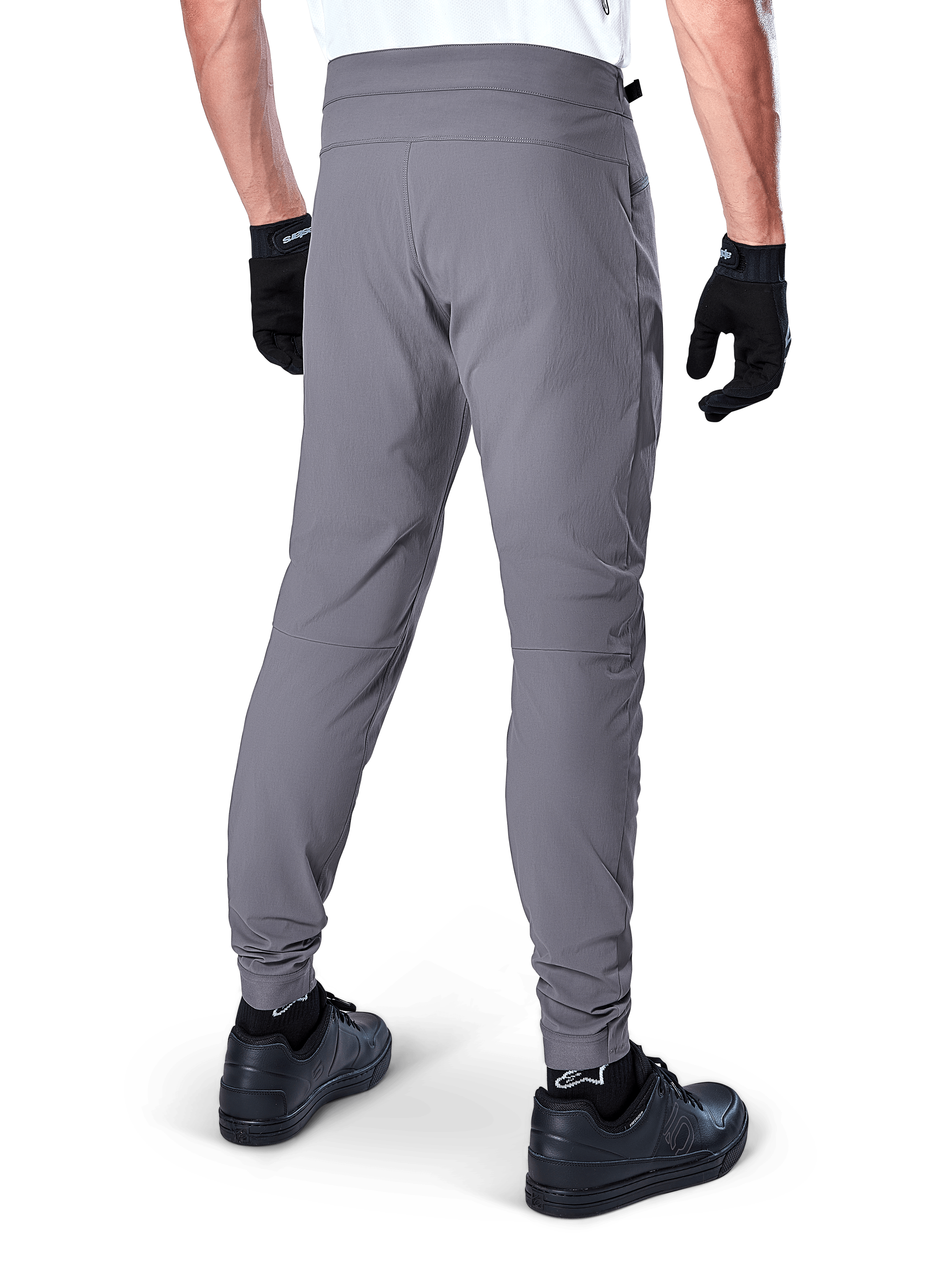 Alpinestars A-Dura Pants, spodnie rowerowe MTB, ciemnoszare, widok z tyłu modelu, wykonane z trwałej dwukierunkowej tkaniny rozciągliwej z elastycznymi mankietami, zestawione z czarnymi rękawiczkami i butami sportowymi dla wyglądu gotowego do jazdy na rowerze