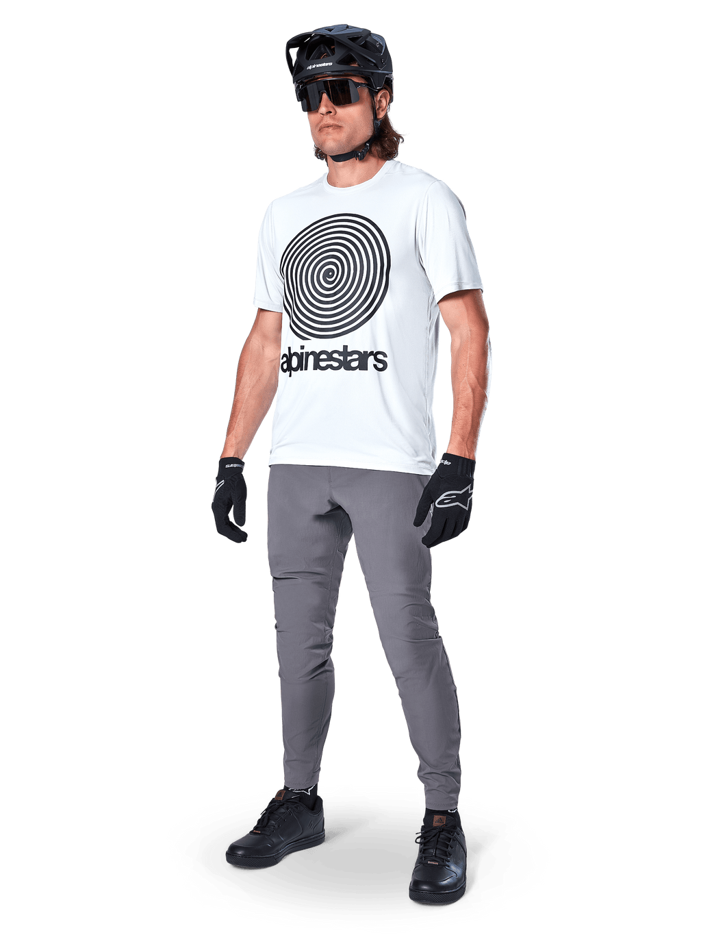 Alpinestars A-Dura Pants, spodnie MTB, ciemnoszare, noszone z białym t-shirtem z grafiką spirali, czarnym kaskiem MTB, okularami przeciwsłonecznymi i czarnymi rękawicami, wytrzymały materiał rozciągliwy w 2 kierunkach do jazdy w terenie