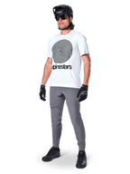 Alpinestars A-Dura Pants, spodnie MTB, ciemnoszare, noszone z białym t-shirtem z grafiką spirali, czarnym kaskiem MTB, okularami przeciwsłonecznymi i czarnymi rękawicami, wytrzymały materiał rozciągliwy w 2 kierunkach do jazdy w terenie