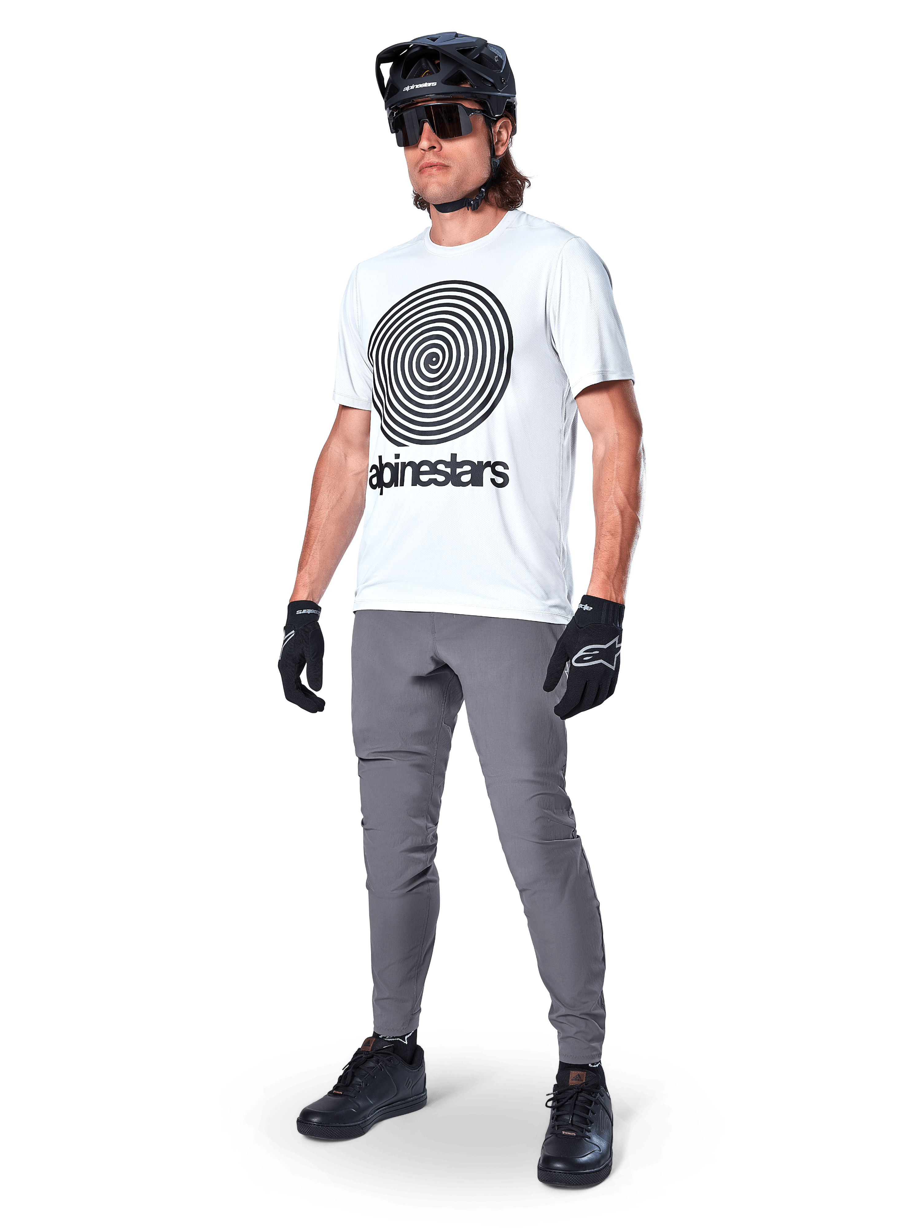 Alpinestars A-Dura Pants, spodnie MTB, ciemnoszare, noszone z białym t-shirtem z grafiką spirali, czarnym kaskiem MTB, okularami przeciwsłonecznymi i czarnymi rękawicami, wytrzymały materiał rozciągliwy w 2 kierunkach do jazdy w terenie