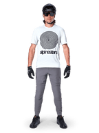 Alpinestars A-Dura Pants, rowerowe spodnie MTB, ciemnoszare, zestawione z białym t-shirtem z grafiką, czarnym kaskiem i rękawiczkami, charakteryzują się trwałą konstrukcją z dwukierunkowego stretchu i regularnym krojem do jazdy na rowerze górskim