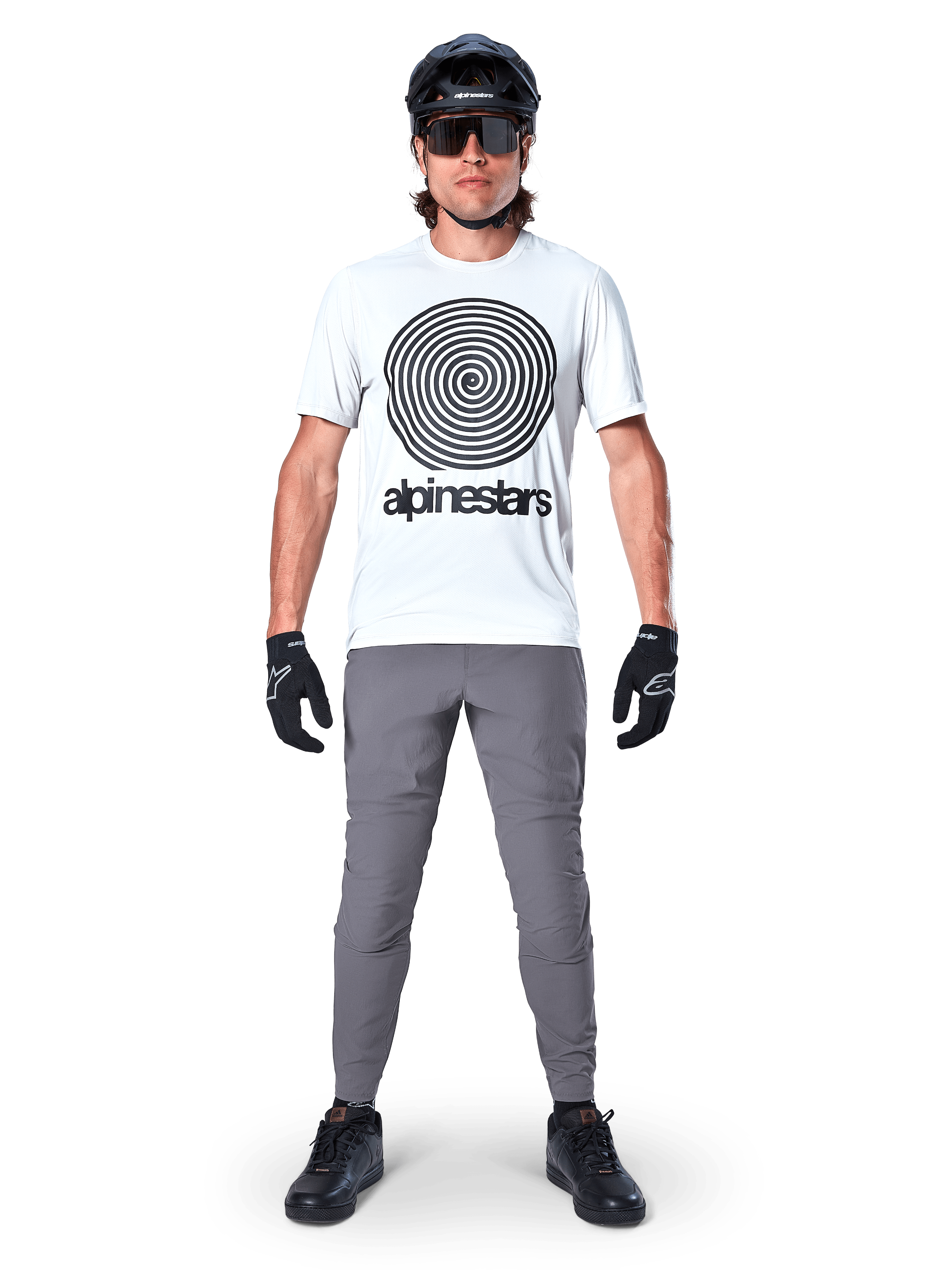 Alpinestars A-Dura Pants, rowerowe spodnie MTB, ciemnoszare, zestawione z białym t-shirtem z grafiką, czarnym kaskiem i rękawiczkami, charakteryzują się trwałą konstrukcją z dwukierunkowego stretchu i regularnym krojem do jazdy na rowerze górskim