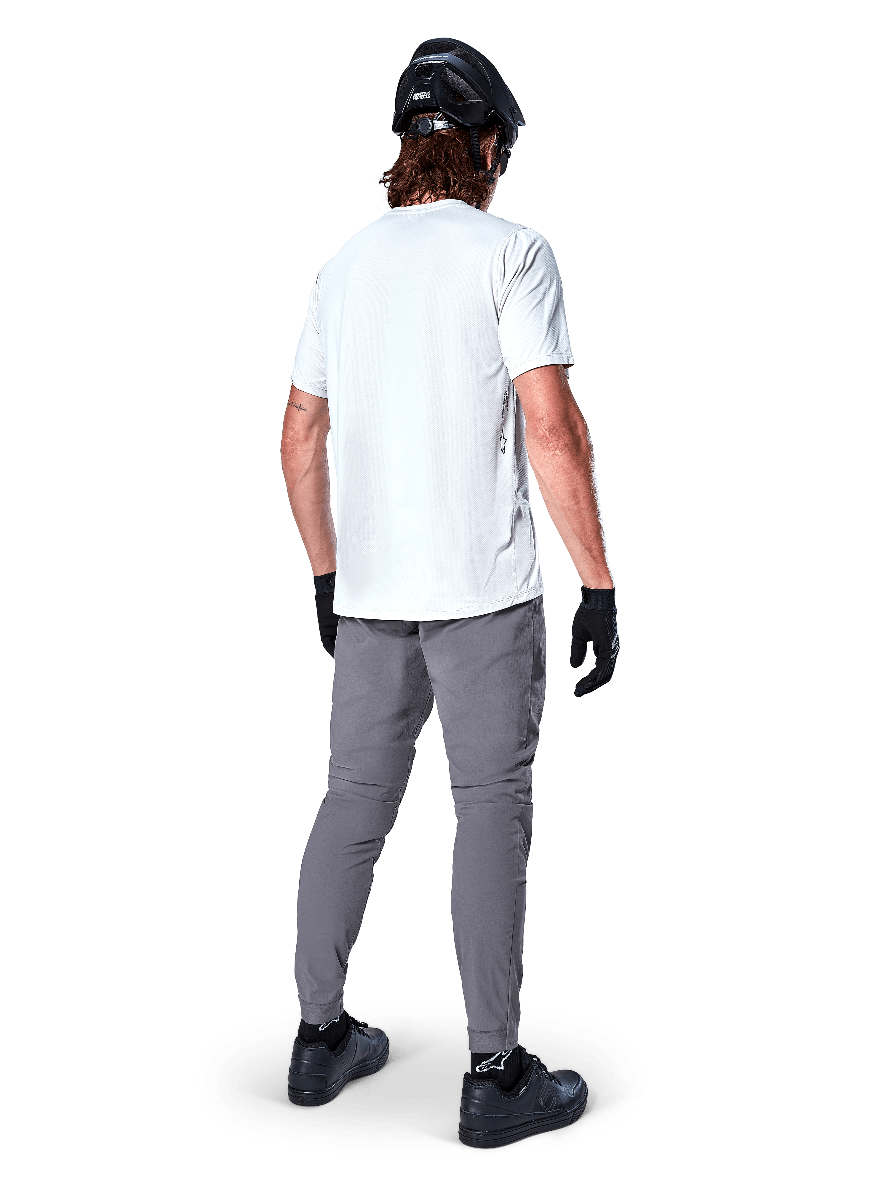 Alpinestars A-Dura Pants, ciemnoszare spodnie na rower górski widoczne z tyłu, zestawione z białym t-shirtem z krótkim rękawem, czarnym kaskiem MTB, czarnymi rękawiczkami i butami, krój regular fit z elastycznymi mankietami, wytrzymała odzież do jazdy w terenie
