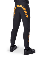 Alpinestars A-Supra Race Pants, czarne spodnie MTB, widok z tyłu z dużym pomarańczowym napisem Alpinestars na karczku, pomarańczowe logo na dolnej części nogawki, sportowy krój z kieszenią na zamek, czarne buty