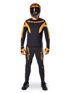 Alpinestars A-Supra Race Pants, spodnie wyścigowe MTB, czarno-pomarańczowe, prezentowane na zawodniku w pełnym stroju wyścigowym z czarno-pomarańczową koszulką, pomarańczowym kaskiem i dopasowanymi rękawicami, z dużymi pomarańczowymi logotypami gwiazdy na nogawkach