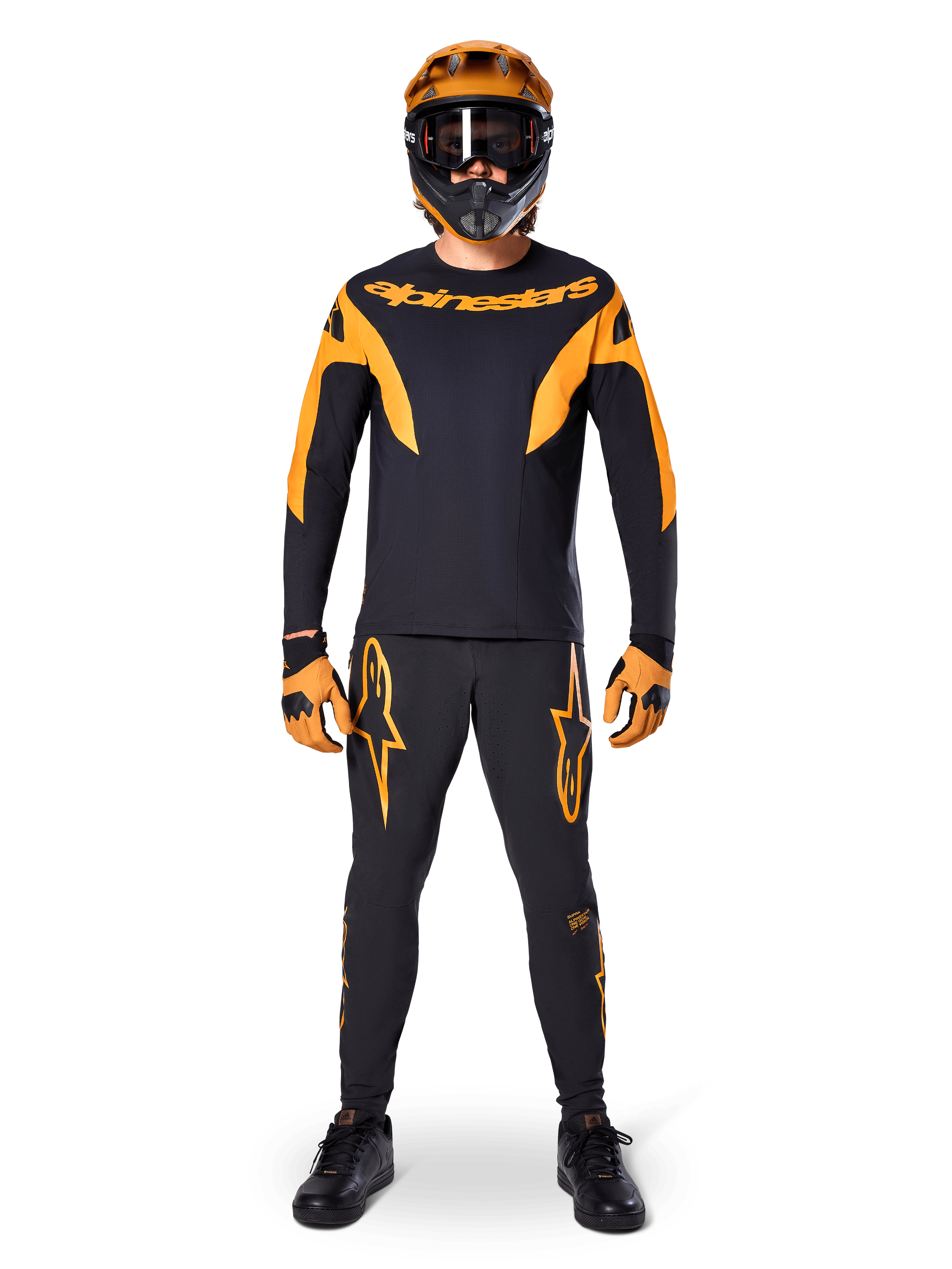 Alpinestars A-Supra Race Pants, spodnie wyścigowe MTB, czarno-pomarańczowe, prezentowane na zawodniku w pełnym stroju wyścigowym z czarno-pomarańczową koszulką, pomarańczowym kaskiem i dopasowanymi rękawicami, z dużymi pomarańczowymi logotypami gwiazdy na nogawkach