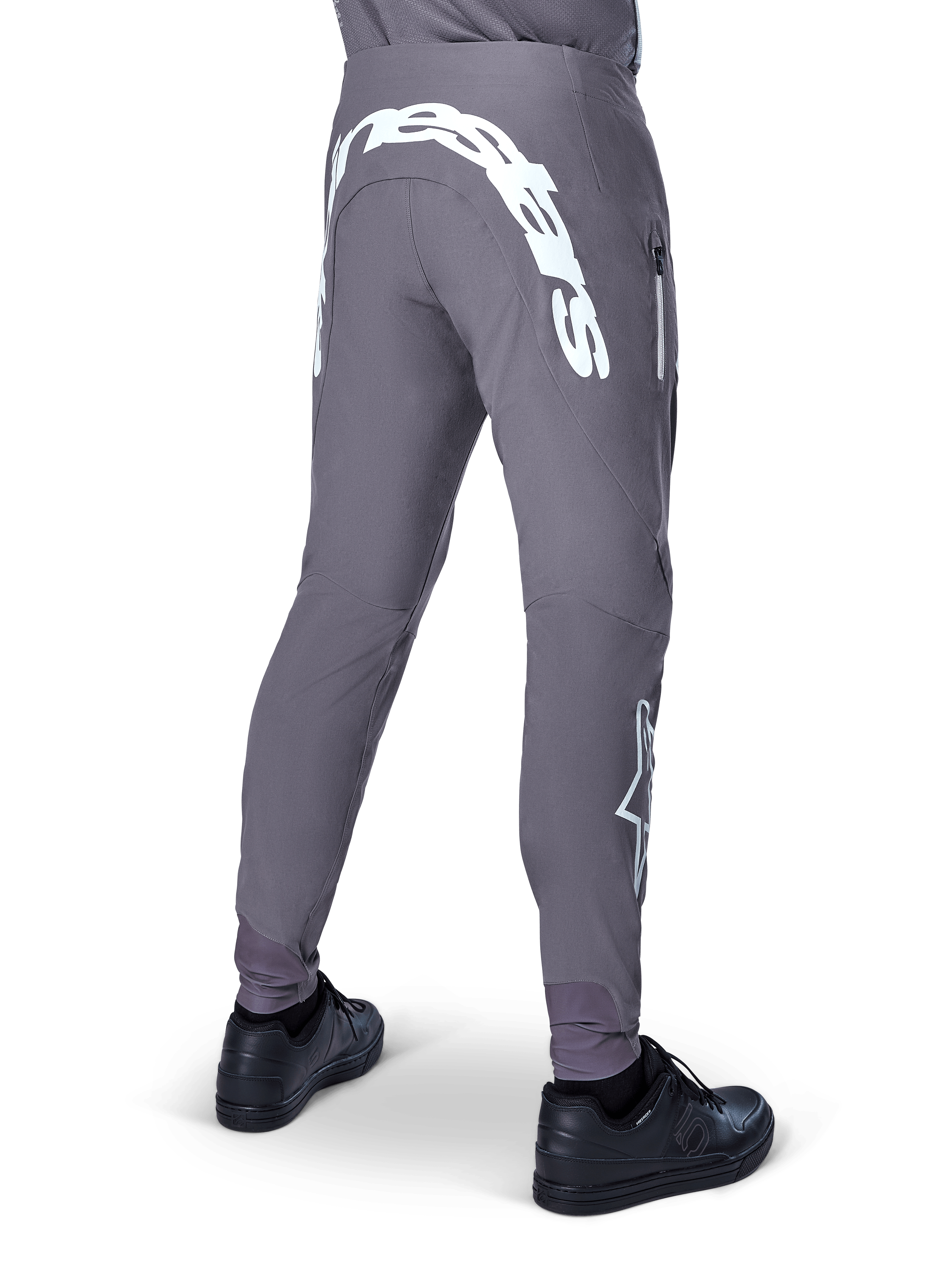 Alpinestars A-Supra Race Pants, spodnie rowerowe MTB, ciemnoszare, widok z tyłu, czarne buty, duży biały napis Alpinestars na karczku i logo gwiazdy na nogawce, kieszeń zapinana na zamek, elastyczny materiał techniczny do wyścigów MTB