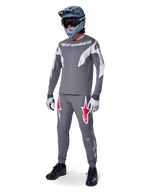 Alpinestars A-Supra Race Pants w kolorze ciemnoszarym, z wyraźnymi czerwonymi i białymi logotypami na udach, noszone przez zawodnika jako część kompletnego zestawu wyścigowego MTB z pasującą szarą koszulką, niebiesko-szarym kaskiem full-face i jasnoszarymi rękawicami, profesjonalna odzież do wyścigów MTB