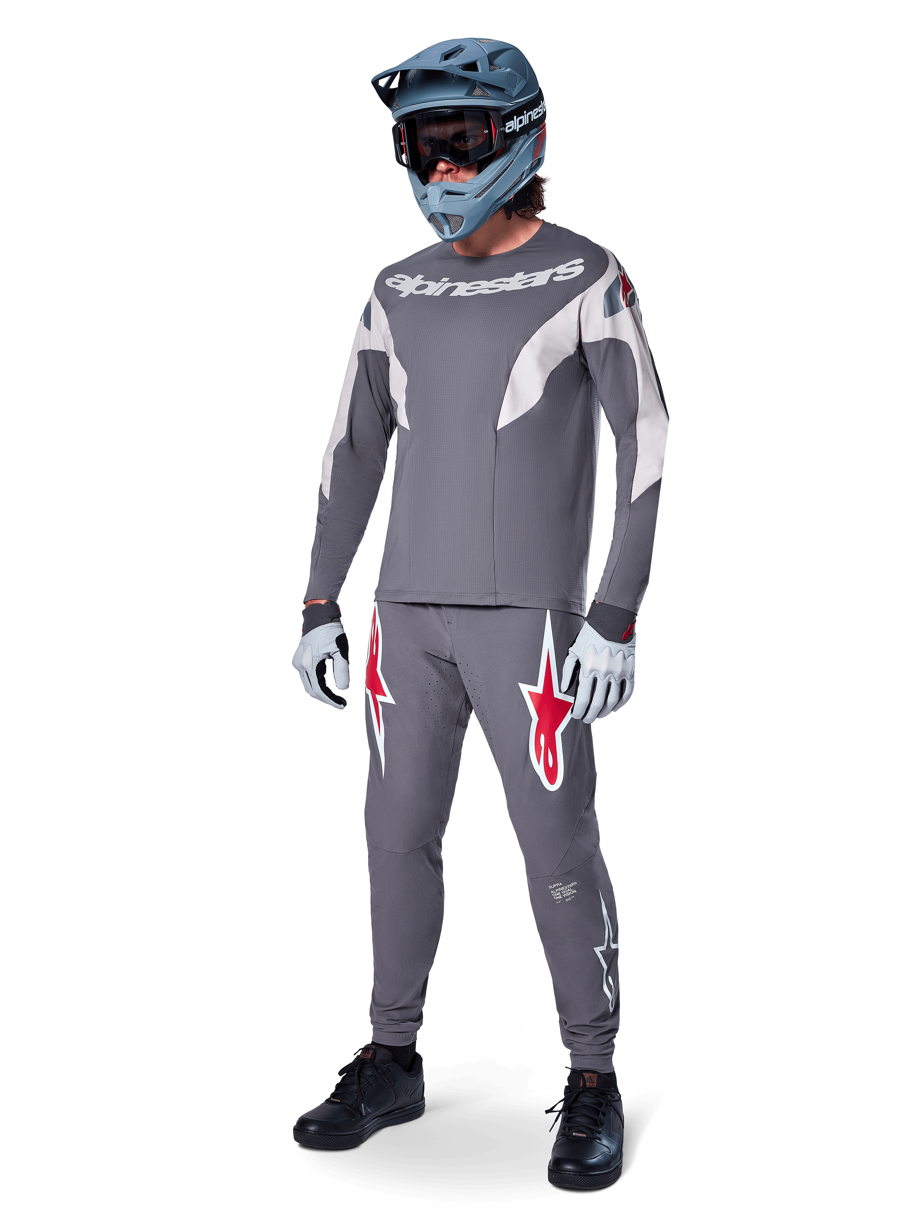 Alpinestars A-Supra Race Pants w kolorze ciemnoszarym, z wyraźnymi czerwonymi i białymi logotypami na udach, noszone przez zawodnika jako część kompletnego zestawu wyścigowego MTB z pasującą szarą koszulką, niebiesko-szarym kaskiem full-face i jasnoszarymi rękawicami, profesjonalna odzież do wyścigów MTB