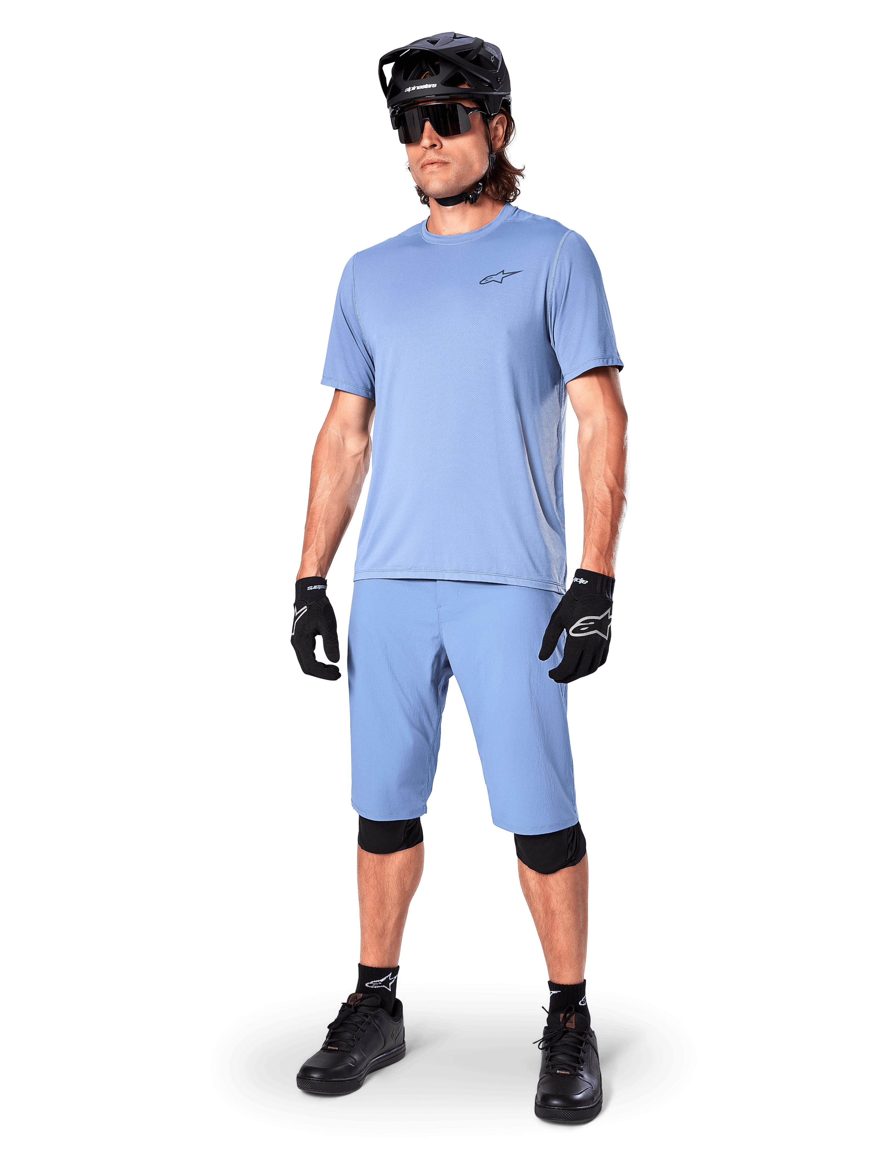 Model ubrany w spodenki Alpinestars A-Dura w kolorze Infinity Blue, pasującą jasnoniebieską koszulkę z krótkim rękawem, czarny kask MTB, okulary przeciwsłoneczne i czarne rękawiczki, kompletny strój do kolarstwa górskiego do jazdy trailowej
