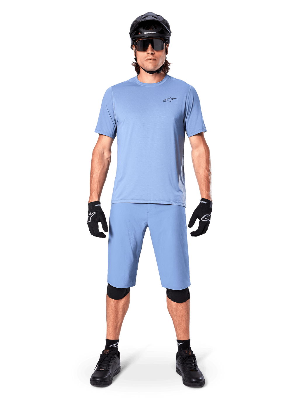 Model w spodenkach Alpinestars A-Dura w kolorze Infinity Blue, pasującej jasnoniebieskiej koszulce, czarnym kasku MTB, czarnych okularach przeciwsłonecznych, czarnych rękawicach i nakolannikach, kompletny strój MTB do jazdy po szlakach