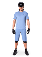 Model w spodenkach Alpinestars A-Dura w kolorze Infinity Blue, pasującej jasnoniebieskiej koszulce, czarnym kasku MTB, czarnych okularach przeciwsłonecznych, czarnych rękawicach i nakolannikach, kompletny strój MTB do jazdy po szlakach