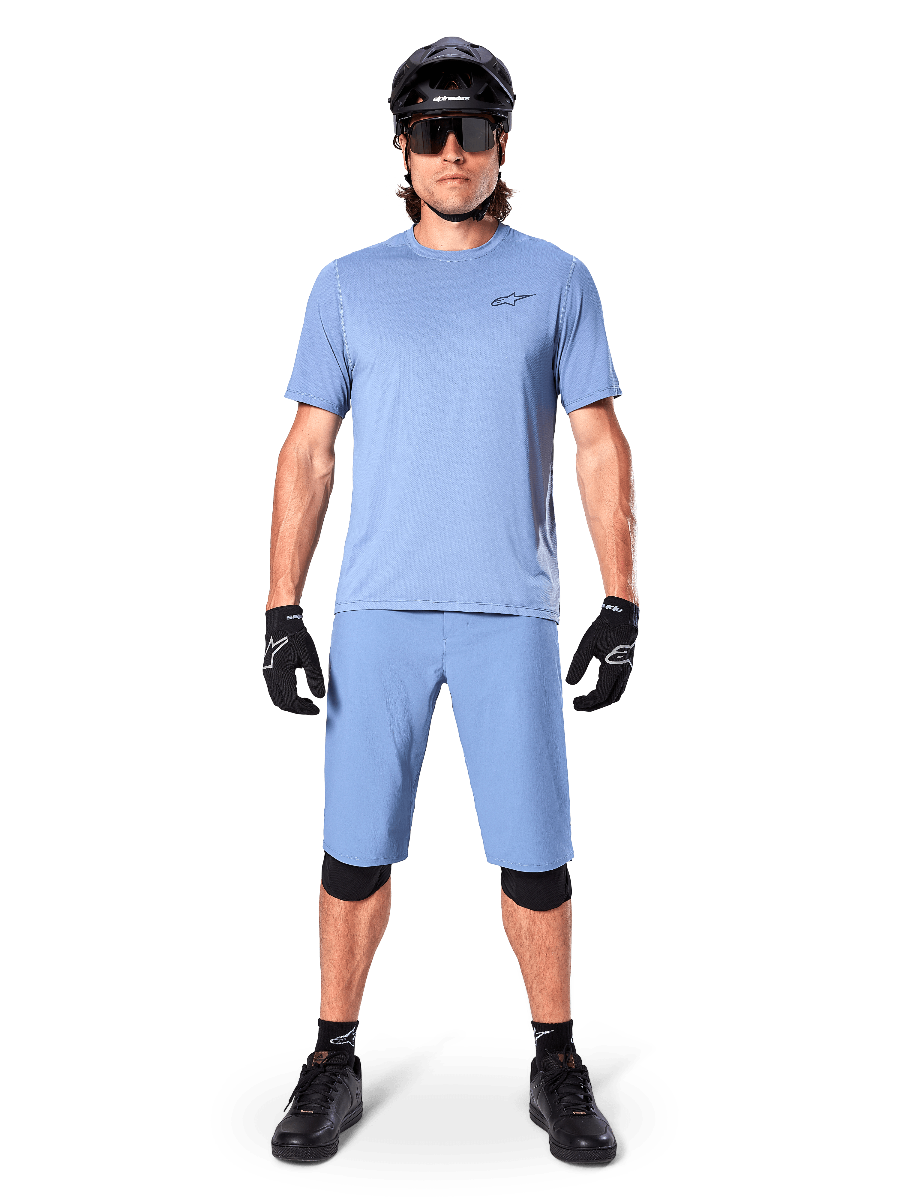 Model w spodenkach Alpinestars A-Dura w kolorze Infinity Blue, pasującej jasnoniebieskiej koszulce, czarnym kasku MTB, czarnych okularach przeciwsłonecznych, czarnych rękawicach i nakolannikach, kompletny strój MTB do jazdy po szlakach
