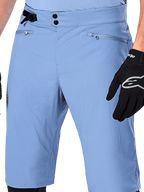 Alpinestars A-Dura Shorts, niebieskie spodenki MTB (Infinity Blue), zbliżenie na talię z podwójnym zapięciem na napy, regulowanym bocznym ściągaczem i kieszeniami na zamek z podszewką z siatki, widoczne z czarną rękawiczką Alpinestars