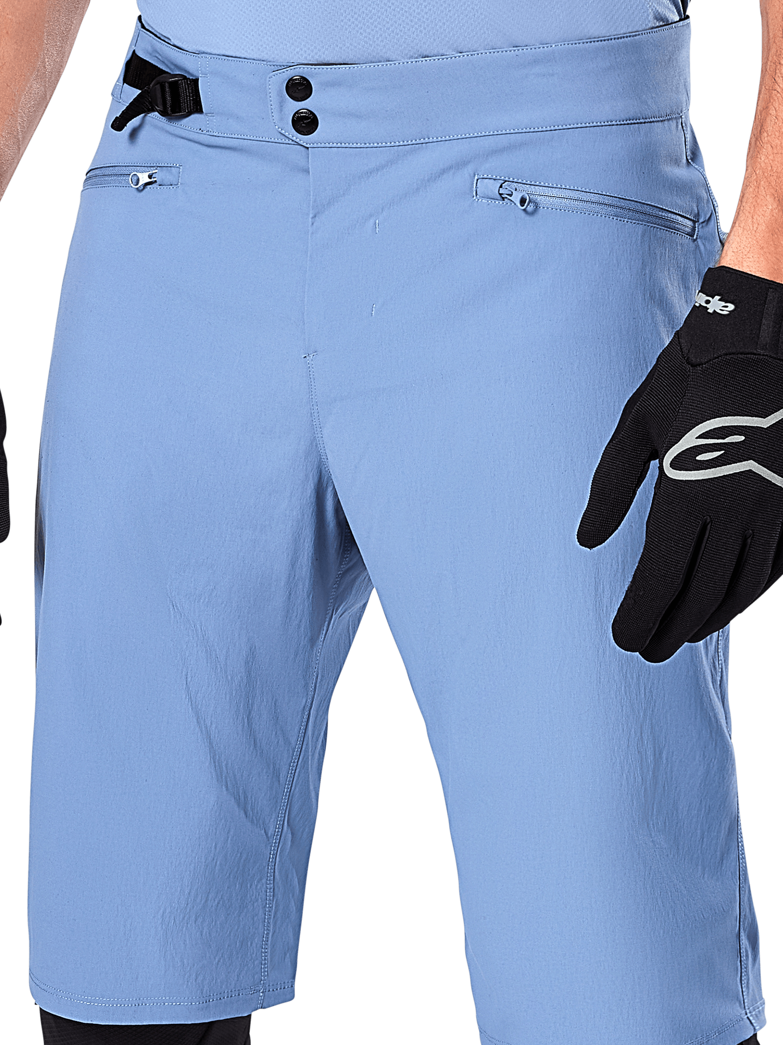 Alpinestars A-Dura Shorts, niebieskie spodenki MTB (Infinity Blue), zbliżenie na talię z podwójnym zapięciem na napy, regulowanym bocznym ściągaczem i kieszeniami na zamek z podszewką z siatki, widoczne z czarną rękawiczką Alpinestars