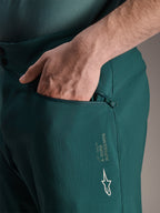 Szorty Alpinestars A-Dura w kolorze Dark Pine, zbliżenie na dłoń w zapinanej na zamek kieszeni z siatkową podszewką, białe logo gwiazdy Alpinestars i branding A-Dura, wytrzymała konstrukcja tekstylna do kolarstwa górskiego