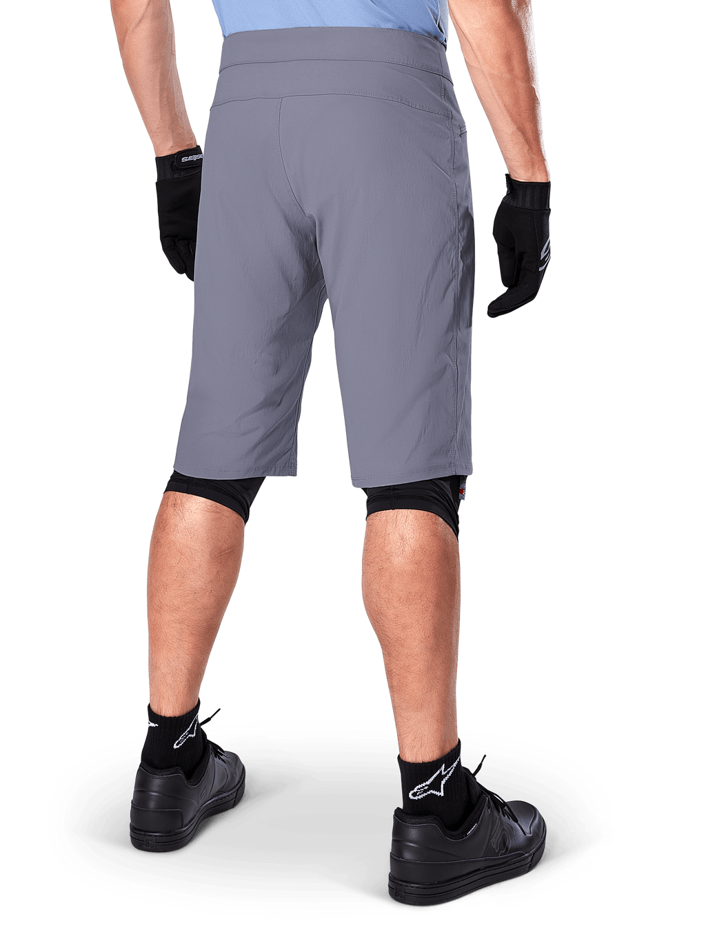 Alpinestars A-Dura Shorts, ciemnoszare spodenki rowerowe MTB, widok z tyłu na rowerzyście, noszone z czarnymi rękawiczkami, czarnymi ochraniaczami kolan i czarnymi skarpetami z logo Alpinestars, wytrzymała konstrukcja do jazdy terenowej
