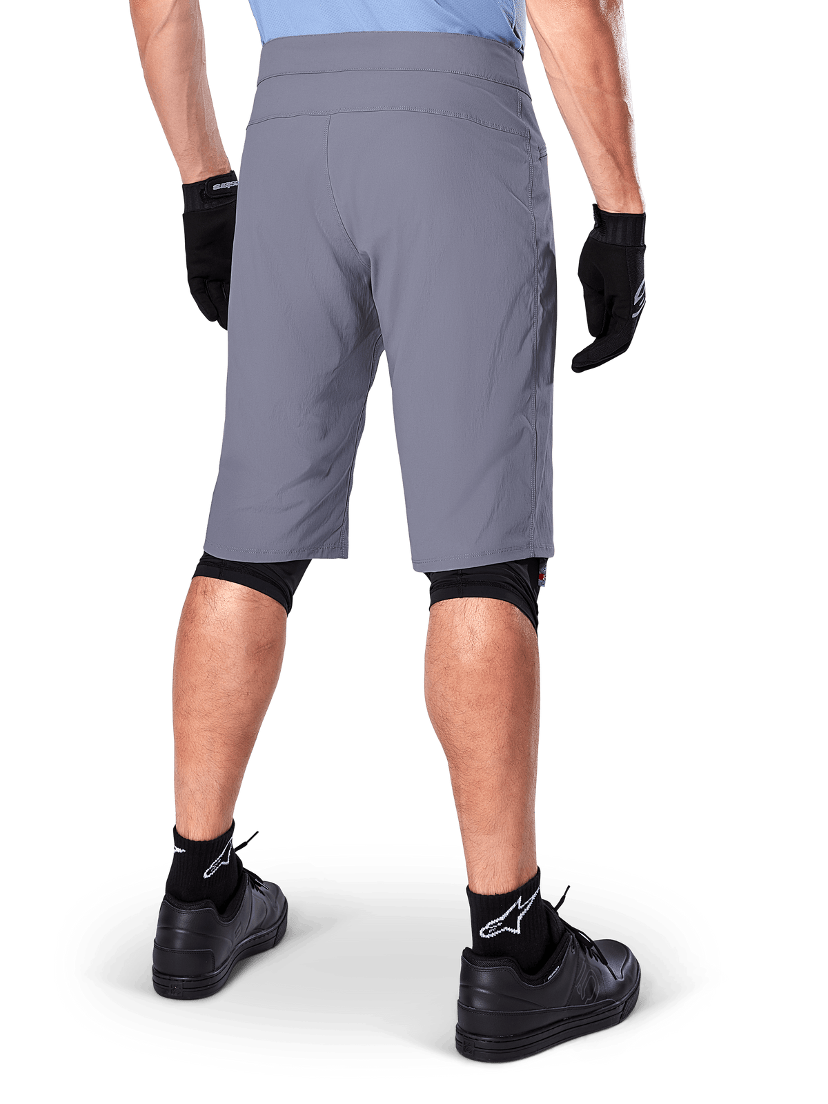 Alpinestars A-Dura Shorts, ciemnoszare spodenki rowerowe MTB, widok z tyłu na rowerzyście, noszone z czarnymi rękawiczkami, czarnymi ochraniaczami kolan i czarnymi skarpetami z logo Alpinestars, wytrzymała konstrukcja do jazdy terenowej