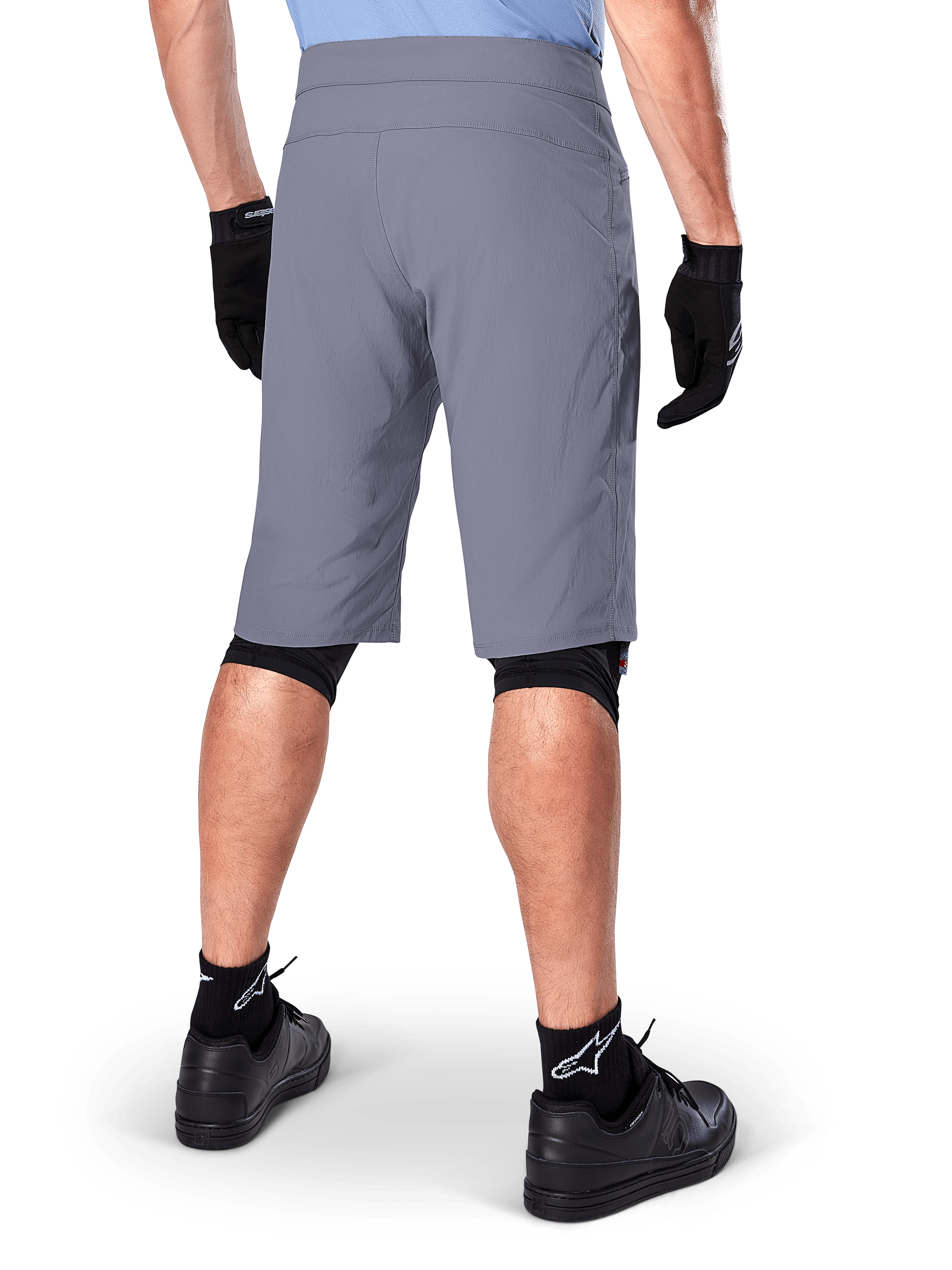 Alpinestars A-Dura Shorts, ciemnoszare spodenki rowerowe MTB, widok z tyłu na rowerzyście, noszone z czarnymi rękawiczkami, czarnymi ochraniaczami kolan i czarnymi skarpetami z logo Alpinestars, wytrzymała konstrukcja do jazdy terenowej