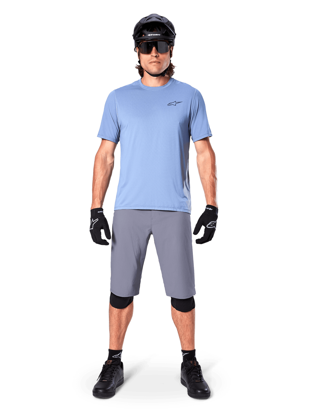 Męski rowerzysta w spodenkach Alpinestars A-Dura w kolorze ciemnoszarym, zestawionych z jasnoniebieskim t-shirtem z krótkim rękawem, czarnym kaskiem, okularami przeciwsłonecznymi i czarnymi ochraniaczami na kolana, kompletny strój do kolarstwa górskiego.