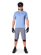 Męski rowerzysta w spodenkach Alpinestars A-Dura w kolorze ciemnoszarym, zestawionych z jasnoniebieskim t-shirtem z krótkim rękawem, czarnym kaskiem, okularami przeciwsłonecznymi i czarnymi ochraniaczami na kolana, kompletny strój do kolarstwa górskiego.