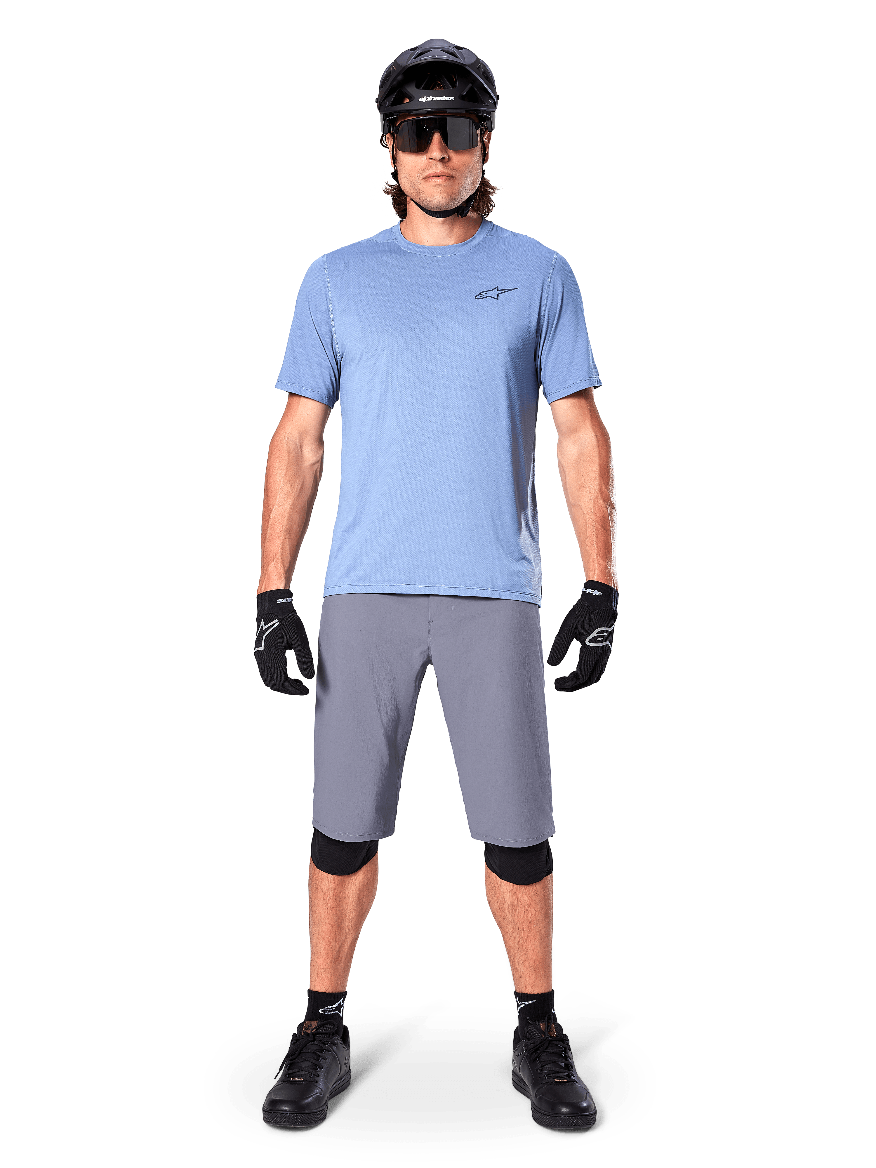 Męski rowerzysta w spodenkach Alpinestars A-Dura w kolorze ciemnoszarym, zestawionych z jasnoniebieskim t-shirtem z krótkim rękawem, czarnym kaskiem, okularami przeciwsłonecznymi i czarnymi ochraniaczami na kolana, kompletny strój do kolarstwa górskiego.