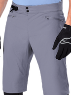 Alpinestars A-Dura Shorts, spodenki MTB, ciemnoszare, zbliżenie na talię pokazujące podwójne zapięcie na napy, boczny ściągacz i kieszenie zapinane na zamek, czarne rękawiczki rowerowe, wytrzymała odzież do kolarstwa górskiego.