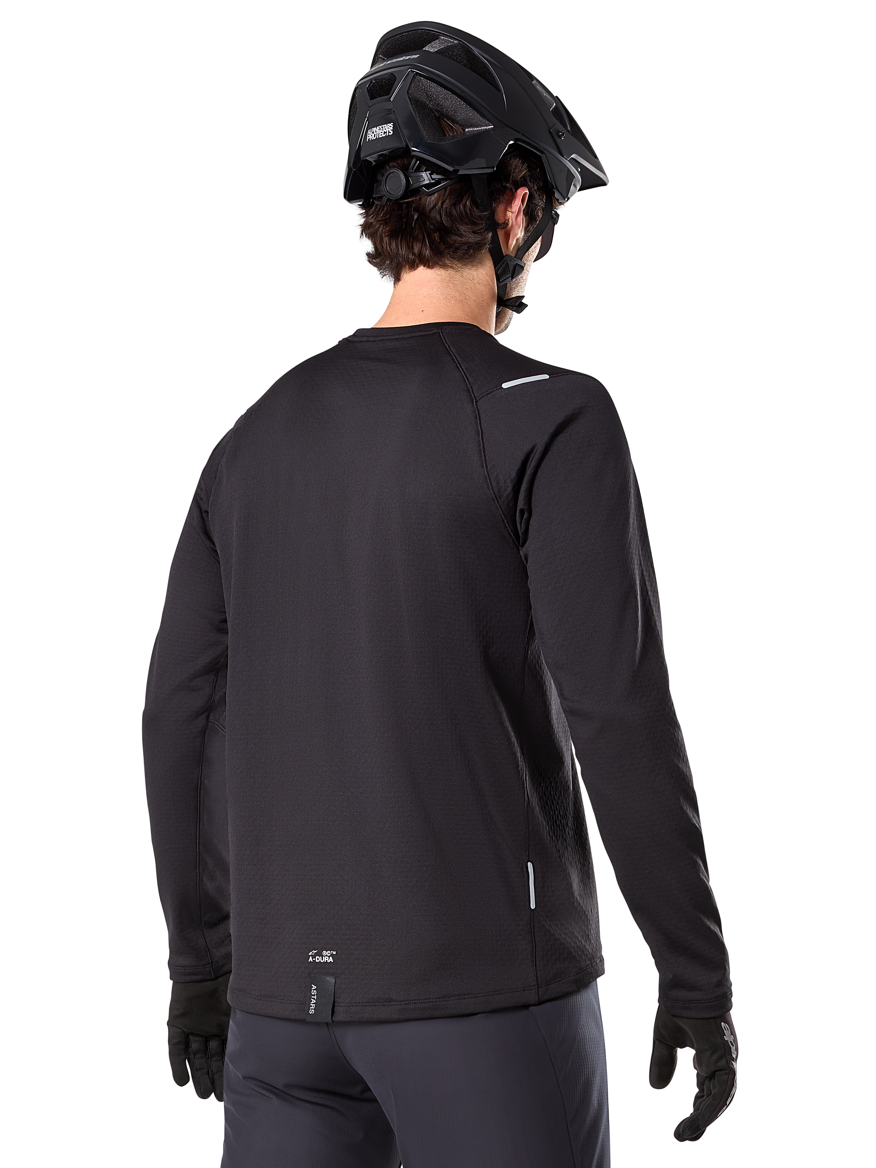 Alpinestars A-Dura Thermal Jersey, koszulka rowerowa MTB z długim rękawem, czarna, widok z tyłu pokazujący przedłużony tył, małe odblaskowe detale i logo na dolnej krawędzi, noszona z czarnym kaskiem i rękawiczkami do jazdy w chłodne dni