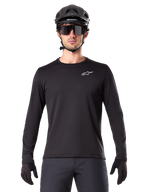 Alpinestars A-Dura Thermal Jersey, czarna koszulka rowerowa MTB z długim rękawem, noszona z czarnym kaskiem i dużymi okularami przeciwsłonecznymi, małe białe logo Alpinestars na piersi, regularny krój, odzież rowerowa MTB