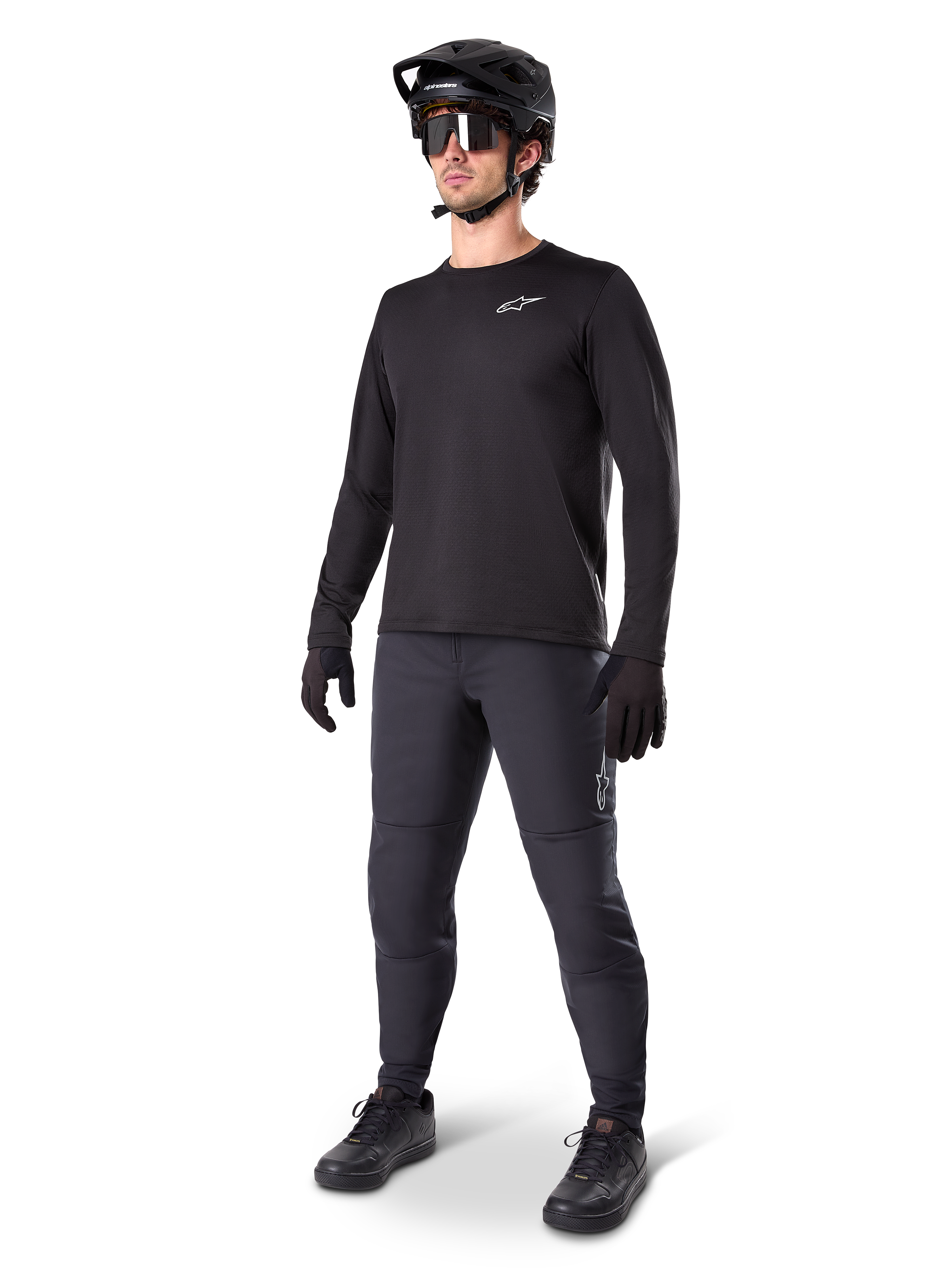 Model w koszulce termicznej Alpinestars A-Dura, czarna koszulka MTB z długim rękawem, czarny kask rowerowy MTB, okulary przeciwsłoneczne i ciemnoszare spodnie, małe białe logo na piersi, izolująca konstrukcja polarowa do jazdy w chłodne dni