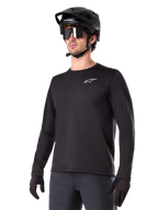 Alpinestars A-Dura Thermal Jersey, czarna koszulka MTB z długim rękawem, białe logo na piersi, noszona z czarnym kaskiem i rękawicami, nowoczesny sportowy design do jazdy na szlaku
